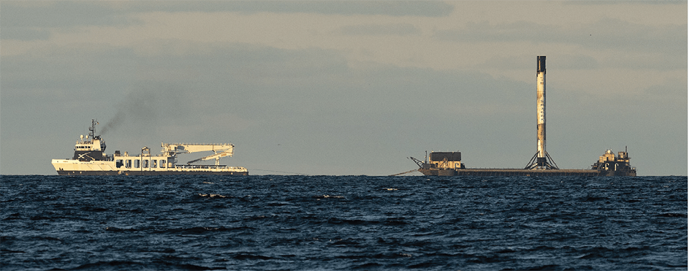A photograph of SpaceX recovery vessel, Doug, moving a SpaceX rocket on a barge through the water.