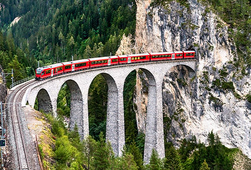 A red train on a stone bridge exiting a tunnel through the side of a mountain in the woods